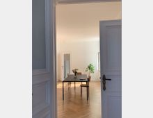 Blick durch eine geöffnete weiße Tür in einen hellen Raum mit Parkettboden. Im Raum stehen ein schwarzer rechteckiger Tisch mit einem Blumenstrauß darauf und vier Stühle; im Hintergrund ist eine große Zimmerpflanze am Fenster zu sehen.