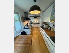 Moderne Küche mit weißen Hochglanzfronten und heller Holzarbeitsplatte auf beiden Seiten eines schmalen Raums. Der Boden besteht aus hellem Holz, und ein Esstisch mit passendem Holzfinish sowie schwarze Lampen mit dekorativen Innenmustern hängen von der Decke.