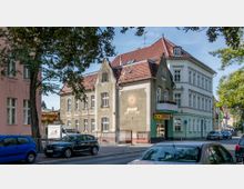 Straßenszene mit einem historischen Gebäude auf der rechten Seite, das ein Schild mit der Jahreszahl „1904“ und einem Sonnenmotiv trägt; daneben ein weiteres Gebäude mit abgerundeten Giebeln und roten Ziegeldächern. Im Vordergrund sind parkende Autos und Bäume zu sehen.