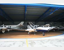 Ein moderner Flugzeughangar mit einer blauen Metallstruktur, der mehrere kleine Propellerflugzeuge beherbergt, darunter ein zweimotoriges Flugzeug im Vordergrund. Der Boden besteht aus Beton- und Holzflächen, und die Flugzeuge sind in geparkter Position.