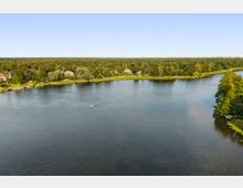 Weitblick über einen breiten See mit ruhiger Wasseroberfläche; am gegenüberliegenden Ufer verläuft ein schmaler Schilfgürtel vor dichtem Wald mit vereinzelten Häusern. Rechts ragt eine bewaldete Landzunge ins Wasser, am Horizont sind in der Ferne Gebäude einer Stadt zu erkennen.