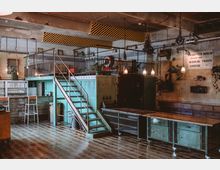 Industrie-inspirierter Raum mit einer Mischung aus rustikalen und modernen Elementen, einschließlich einer Metalltreppe zu einer erhöhten Fläche mit Geländer, hölzernem Boden, grünen Schränken und Hängelampen. An der Wand sind alte Schilder und Pflanzen angebracht, was dem Raum ein Vintage-Flair verleiht.