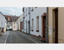 Schmale, gepflasterte Altstadtstraße mit weißen und beige-braunen Gebäuden, teils mit Fachwerkelementen. Straßenlaternen und Briefkästen sind sichtbar, im Hintergrund steht ein geparktes Auto.