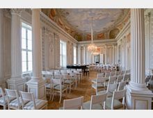 Eleganter, barocker Veranstaltungssaal mit dekorativen weißen Säulen, aufwändig verzierten Wänden und einer Decke mit Wolkenfresko. Der Raum ist mit weißen Stühlen und einem schwarzen Flügel ausgestattet, ein großer Kronleuchter hängt zentral von der Decke.