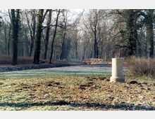Waldpark mit kahlen Bäumen im Winter, einem kleinen Holzsteg über einen Wassergraben und einer steinernen Säule im Vordergrund. Der Boden ist teilweise mit Laub bedeckt und von Frost überzogen.