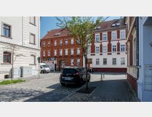 Straßenszene in einer Wohngegend mit gepflastertem Boden, einem geparkten schwarzen Auto und einem Baum im Vordergrund. Im Hintergrund stehen historische Gebäude mit roten Backsteinfassaden und weißen Fensterrahmen.