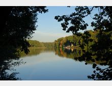 Ein ruhiger See, umgeben von dicht bewaldeten Ufern, mit einer kleinen gelben Gebäudeanlage und Bootsanlegern im Hintergrund. Überhängende Äste und Blätter rahmen den Blick auf das Wasser ein.