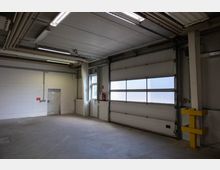 Leerer Lagerraum mit Betonboden, einer weißen Tür in der Rückwand sowie einem großen Sektionaltor mit halbtransparenten Fenstern auf der rechten Seite. Belüftungs- und Rohrleitungen verlaufen an der Decke, und eine gelbe Sicherheitsbarriere ist nahe der Wand sichtbar.