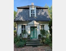 Kleines Einfamilienhaus mit weißer Fassade und dunkel gedecktem Satteldach, zentraler, grüner Eingangstür mit dekorativem Kranz sowie einem kleinen Treppenaufgang. Umgeben von einem gepflegten Garten mit Blumen und Sträuchern.