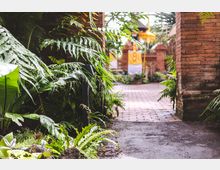 Schattiger Gartenpfad mit üppigen grünen Farnen und tropischen Pflanzen, flankiert von roten Backsteinmauern und -säulen; im Hintergrund ein kleiner Pavillon mit gelber und weißer Dekoration.
