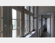 Ein Flur in einem Gebäude mit weißen Fliesen an den Wänden, großen Fenstern mit Sprossenrahmen auf der linken Seite und weißen Türen, die zu angrenzenden Räumen führen. Auf der Fensterbank steht eine kleine Vase mit Blumen und eine braune Flasche.