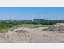 Offene Landschaft mit Windkraftanlagen im Hintergrund, umgeben von Feldern, Wäldern und sanften Hügeln. Im Vordergrund sind Erdhaufen und Schotter mit spärlicher Vegetation zu sehen.