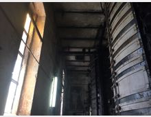 Ein Industrieinnenraum mit hohen, verwitterten Wänden und einem schmalen Fenster, durch das Tageslicht fällt. Rechts im Bild befinden sich große, vertikale Metallstrukturen mit mehreren übereinander liegenden Verschlüssen.