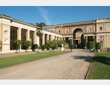 Historisches Gebäude mit klassizistischer Architektur, gekennzeichnet durch große Fenster, Säulen und Verzierungen. Vor dem Gebäude befinden sich gepflegte Grünflächen, Topfpflanzen und ein zentraler Weg, der zu einem Torbogen führt.