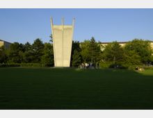 Eine Beton-Skulptur mit drei vertikalen Spitzen steht auf einer großen grünen Rasenfläche, umgeben von Bäumen und einem hellen Gebäude im Hintergrund. Eine Gruppe von Menschen steht nahe der Skulptur, und weitere Personen sitzen im Gras.