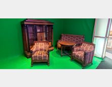 Ein Raum mit grünem Hintergrund, eingerichtet mit antiken Holzmöbeln, darunter zwei gepolsterte Sessel, ein Sofa, ein runder Holztisch und ein großer Vitrinenschrank. Rechts ist ein Bereich mit einer teils durchsichtigen Wand sichtbar.