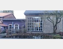 Backsteingebäude mit einer großen Fensterfront und angrenzendem Flachbau mit lila verputzter Fassade. Im Vordergrund befinden sich ein kleiner Teich, ein Holzzaun sowie ein Baum ohne Laub.