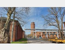 Industriegelände mit roten Backsteingebäuden, einem markanten Wasserturm im Hintergrund und metallischen Rohrleitungen, die zwischen den Gebäuden verlaufen; im Vordergrund stehen Bäume und ein Geländewagen.