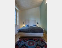 Schlafzimmer mit Holzboden und -decke, einem Doppelbett mit grauer Bettdecke und blauen Kissen. Vor dem Bett liegt ein farbenfroher Teppich mit traditionellen Mustern, und die Wand links hat ein großes Fensterrahmen aus Holz.