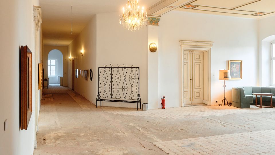 Großer, heller Flur in einem historischen Gebäude mit Kronleuchter, dekorierter Decke und hohen Doppeltüren. Links führt ein langer Korridor zu einem Bogenfenster; rechts stehen eine Sitznische mit Sofa, Tisch und Stehlampe neben einem großen Fenster.