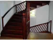 Holztreppe mit dunklem Holzgeländer und Gittermuster-Details, die sich in einem hellen Treppenhaus mit weiß gestrichenen Wänden befindet. Im Hintergrund ist ein Fenster, das Tageslicht hereinlässt.