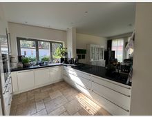 Moderne Küche mit weißer Einbauküche und schwarzen Arbeitsplatten, großen Fenstern, die Tageslicht hereinlassen, und einem hell gefliesten Boden. Im Hintergrund sind ein Wohnzimmerbereich mit Glaskommoden und Vorhängen zu sehen.