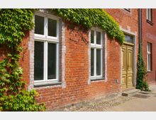 Rote Ziegelhausfassade mit großen weißen Fenstern, einer Holztür in warmem Braunton und grünem Efeu, der einen Teil der oberen Wand bedeckt. Der Boden vor dem Gebäude ist mit Kopfsteinpflaster und Gehwegplatten gestaltet.