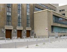 Großes Gebäude mit einer Fassade aus grauem Stein und mehreren hohen Fenstern. Der Eingangsbereich zeigt drei massive Holztüren, flankiert von Außenleuchten, während die angrenzenden Bereiche modernere Strukturen mit Glasfenstern und Betonwänden aufweisen.