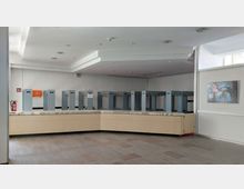 Heller Raum mit einer Garderobe, bestehend aus grauen Metallständern mit numerierten Hakenreihen. Im Vordergrund eine lange Empfangstheke aus hellem Holz, an der rechten Wand hängt ein Gemälde, und eine Feuerlöscherstation ist an der linken Wand angebracht.