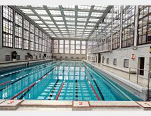 Hallenbad mit einem langen Schwimmbecken, das durch rot-weiße Abtrennungsleinen in Bahnen unterteilt ist. Die Wände und das Dach bestehen aus großen Glasfenstern, die viel Licht hereinlassen; seitlich befinden sich Sitzbänke und Notausrüstungen.