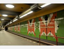 U-Bahn-Station Mierendorffplatz mit einer langen, leicht gebogenen Plattform. Eine Wand mit grün-rotem Mosaikmuster und Schriftzug „Mierendorffplatz“ dominiert die Szene, flankiert von einer gelben Decke mit eingebauten Leuchtkörpern.
