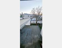 Blick von einer erhöhten Terrasse auf eine Reihenhaussiedlung mit flachen, weißen Fassaden, einem kahlen Baum im Garten und einem klaren Himmel im Hintergrund. Im Vordergrund ist ein Flachdach sichtbar.