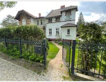 Zweistöckiges Wohnhaus mit weißer Fassade und roten Backsteindetails, umgeben von einem gepflegten Garten. Im Vordergrund ein schwarzer Metallzaun mit einem Weg, der zum Hauseingang führt.