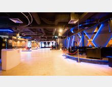 Ein moderner Innenraum einer Veranstaltungs- oder Club-Lounge mit einer offenen Fläche, schwarzen Sitzmöbeln, roten Abgrenzungsseilen und blauen LED-Lichteffekten an den Wänden. An der Decke befinden sich sichtbare Rohre und runde Lichtinstallationen.