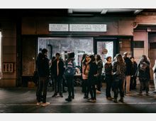 Eine Gruppe von Menschen steht vor einem beleuchteten Gebäude in einer Stadtstraße bei Nacht. Über dem Eingang des Gebäudes hängt ein Schild mit der Aufschrift „BI BAX CHANGE #17 DAUGHTERS & SONS OF GASTARBEITERS 20:00“, während im Hintergrund ein industriell gestalteter Innenraum zu sehen ist.