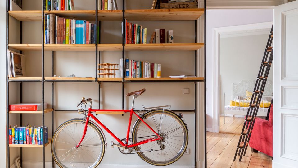 Heller Wohnraum mit großem, offenen Bücherregal aus Holz und schwarzem Metall, gefüllt mit Büchern und einigen Gegenständen; darunter steht ein rotes Fahrrad vor der Regalwand. Rechts führt eine offene Tür in ein Schlafzimmer mit Metallbett, daneben lehnt eine schwarze Leiter am Durchgang.