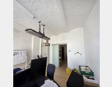 Ein einfacher Büroraum mit weiß gestrichenen Wänden und Holzdielenboden. Zu sehen sind Schreibtischstühle, Computerbildschirme, ein Whiteboard mit angehängten Papieren, eine Hängelampe an Ketten, Regale im Hintergrund sowie gelbe und grüne Kisten mit Dokumenten.