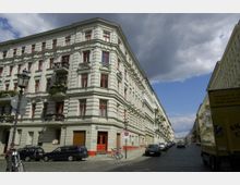 Ecke eines historischen Wohngebäudes im Stil des 19. Jahrhunderts mit heller Fassade, roten Fensterrahmen und Balkonen mit Blumen, gelegen an einer gepflasterten Straße. Parkende Autos und ein gelber Lieferwagen befinden sich am Straßenrand, während im Hintergrund ein leicht bewölkter Himmel zu sehen ist.