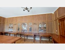 Holzvertäfelter Raum mit dekorativen Intarsien an den Wänden, die figürliche und naturbezogene Motive zeigen. Der Raum ist mit langen Holztischen und einem Kronleuchter an der Decke ausgestattet.