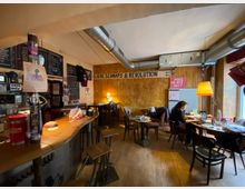 Innenraum einer gemütlichen Bar oder eines Cafés mit langer Theke links, Holzwand und mehreren kleinen Tischen und Stühlen auf Holzboden. Sichtbare Lüftungsrohre an der Decke, Wandtafeln mit Speisekarte und politische Poster, dazu warme Lampenbeleuchtung.