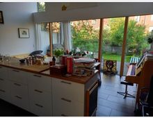 Wohnbereich mit offener Küche und großen Fenstern, die auf einen Garten mit Rasen und Bäumen blicken. Im Vordergrund eine Kücheninsel mit Holztischplatte, im Hintergrund ein Klavier und ein dekoratives Glasbild vor der Terrassentür.