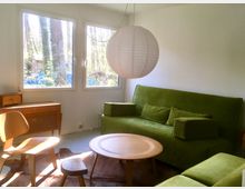 Wohnzimmer mit minimalistischer Einrichtung: Eine grüne Couch, ein runder Holztisch und ein Holzhocker stehen im Raum, beleuchtet von großen Fenstern, die auf eine bewaldete Außenszene blicken. Eine runde Papierlampe hängt von der Decke, daneben ein Holzstuhl und ein Sideboard.