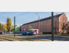 Straßenszene mit einer Straßenbahn-Haltestelle, mehrstöckigen orangefarbenen Wohngebäuden und geparkten Autos im Vordergrund; daneben befinden sich vereinzelte Bäume und Strommasten.