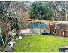 Ein Garten mit Rasenfläche, umgeben von Bäumen und Sträuchern, mit einer Holzpergola im Hintergrund, die farbige Glaselemente mit gemalten Motiven enthält. Links steht eine steinerne Engelstatue, daneben liegen Holzscheite gestapelt, und rechts sind Holzverschläge und Gartengeräte zu sehen.