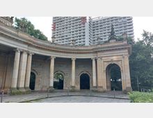 Historischer Kolonnadenbau mit Säulen und rundbogigen Durchgängen, verziert mit Reliefs, im Vordergrund einer gepflasterten Fläche; im Hintergrund moderne Hochhäuser und Bäume.