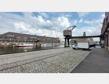 Industriehafen mit historischen Backsteingebäuden entlang eines Kanals, links liegen Boote am Wasser, in der Mitte ein alter Industriekran mit Metallsteg. Im Vordergrund ist eine gepflasterte Fläche mit Schienen, ein geparktes Lieferfahrzeug steht rechts.