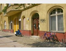 Straßenansicht eines Wohnhauses mit gelblich-beiger Fassade und roten Fensterumrandungen, verziert mit Rundbogenfenstern. Vor dem Gebäude stehen ein blau-weißer Motorroller und ein blaues Fahrrad auf einem gepflasterten Gehweg; daneben ist eine holzverzierte Eingangstür und Graffiti an der Wand sichtbar.