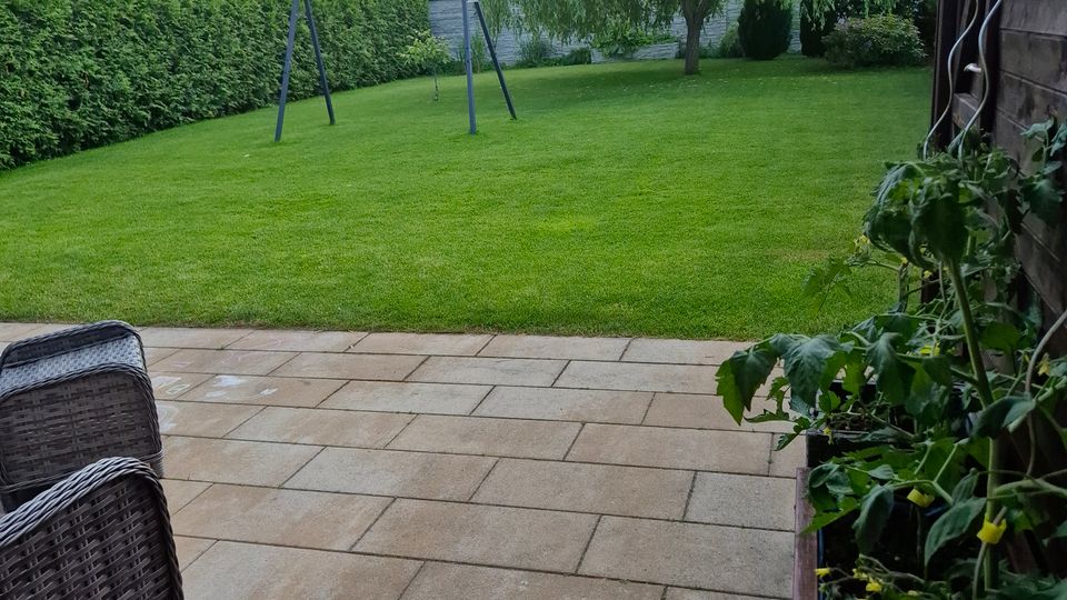 Blick von einer gepflasterten Terrasse in einen großen, gepflegten Garten mit Rasenfläche, hoher Hecke am Rand und einer einzelnen Schaukel. Im Hintergrund steht ein großer, herabhängender Baum, rechts ist die Außenwand eines Holzgebäudes mit Pflanzkübel zu sehen.
