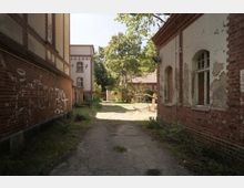 Ein verlassener Innenhof zwischen alten Backsteingebäuden mit abblätternder Farbe und Graffiti an den Wänden. Im Hintergrund sind Bäume und ein weiteres Gebäude mit roten Ziegeln und einem Ziegeldach zu sehen.