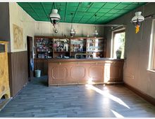 Ein kleiner, rustikaler Barraum mit einer Holztresenfront und einer Regalwand mit Gläsern und Getränkeflaschen. Der Raum hat eine grüne Decke, Hängelampen im Retro-Stil, Holzboden und große Fenster, die natürliches Licht hereinlassen.